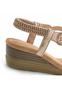 Keilabsatzsandale mit bronzefarbenem Metallic-Finish, elastischen Riemendetails, dekorativen Edelsteinakzenten sowie einem strukturierten, hellbraunen Fußbett und einer dunkelbraunen Laufsohle.