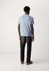 Ljusblå pikétröja med korta ärmar och texturerad yta, kombinerad med mörka jeans och bruna slip-in-skor, sedd bakifrån.