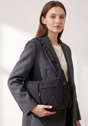 Donna con cappotto grigio e top bianco tiene una borsa a tracolla nera con molte tasche con cerniera e una tracolla con fantasia.