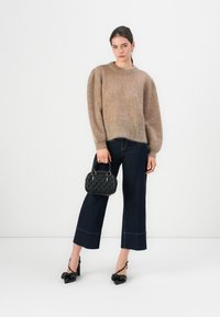 Donna che indossa un maglione beige con maniche a sbuffo, jeans scuri a vita bassa, tacchi a fiocco neri, mentre tiene una borsa nera texture su uno sfondo bianco.