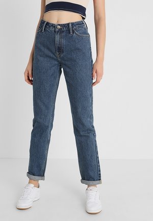 Persoon draagt opgerolde blauwe spijkerbroek, witte sneakers en een cropped beige top met een marineblauwe streep, staand tegen een egale lichte achtergrond.