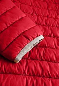 Mouw van een rode gewatteerde jas met horizontale stiksels en een beige elastische boorden, zichtbaar tegen de bijpassende rode gewatteerde stoffen achtergrond.