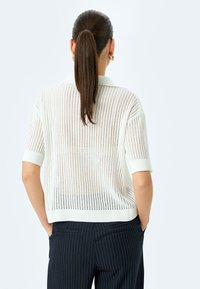 Maglietta polo bianca lavorata a maglia con maniche corte, caratterizzata da un motivo a rete. Abbinata a pantaloni scuri a righe sottili. La vista posteriore evidenzia la texture.