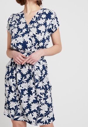 Femme portant une robe bleu marine avec des motifs floraux blancs, manches courtes, et un col en V, debout les mains jointes à la taille.