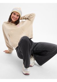 Beige cropped hoodie met een voorzak, grijze wijde spijkerbroek en witte sneakers met rode accenten. Model draagt een meerkleurige gebreide beanie.