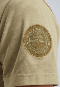 Beige kortærmet skjorte med ribbet tekstur. Har en cirkulær broderet lap på skulderen med tekst og et grafisk design.