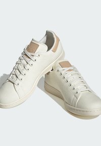 Hvide lærredssneakers med en struktureret overdel, beige læderdetaljer på hælen og flade hvide snørebånd. Rund tå og gummisål.
