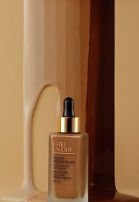Suero de tinte para piel Futurist de Estee Lauder en un frasco de vidrio con tapa dorada y gotero. El fondo presenta texturas fluidas de tonos marrón y beige.