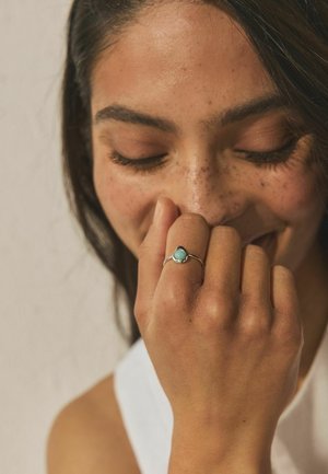 Young woman with closed eyes and freckles lightly touching her face, wearing a silver ring with a green stone on her finger.