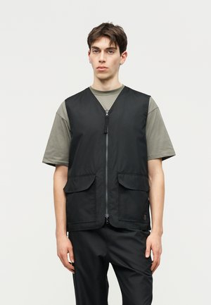 Noor mees, kes kannab musta lukuga vestiga fläpp taskutega oliivirohelise lühikese varrukaga särgi peal, seisab ühevärvilise heleda tausta ees.