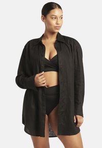 Schwarzes Leinenhemd mit Knopfverschluss, lockerer Schnitt und langen Ärmeln, über einem schwarzen Bikini-Oberteil und hochgeschnittenem Höschen getragen.