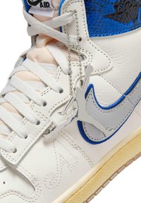 Hvid og blå højtop-sneaker med hvide snørebånd og sølvfarvet Jumpman-logo-nøglering hængende nær Nike-swooshen.