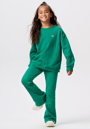 Une fille souriante avec de longs cheveux porte un sweat vert et un pantalon assorti, équilibrée sur un pied devant un fond blanc uni.