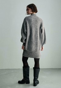 Abito maglione grigio oversize con texture a costine, abbinato a stivali neri alti fino al ginocchio con accessori in argento e tacchi robusti.