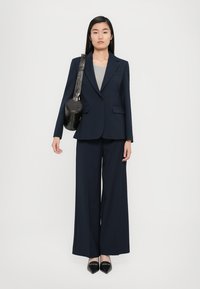 Completo blu navy con blazer sartoriale e pantaloni a gamba larga. Indossato con un top grigio e una borsa rotonda nera con tracolla strutturata. Scarpe nere appuntite.