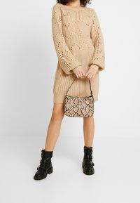 Abito beige in maglia con maniche morbide, abbinato a una piccola borsa a tracolla in stampa pitone marrone e stivaletti neri alla caviglia con fibbie.