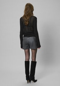 Schwarzes, durchsichtiges Langarmshirt, graue Shorts und schwarze kniehohe Stiefel. Glatte Textur mit dezenten Passformdetails. Rückansicht vor einem grauen Hintergrund.