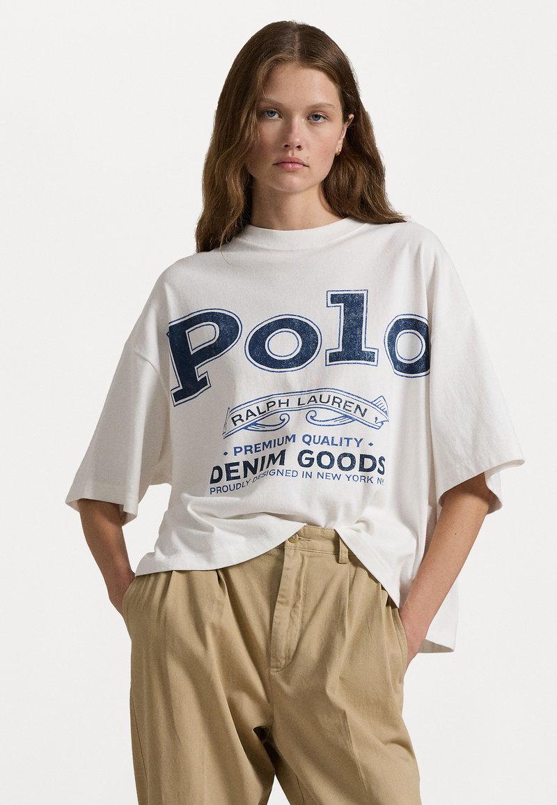 Bílá oversized tričko s modrou grafikou "Polo" a potiskem. Krátké rukávy, kulatý výstřih a volný střih. U páru s béžovými kalhotami.