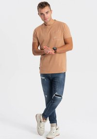 Lysbrun t-shirt med korte ærmer og lomme, kombineret med slidte blå jeans og beige høj-top sneakers. Glat stof, afslappet design.