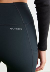 Närbild av en person som bär svarta Columbia-tights och en svart ribbad långärmad tröja, som visar sid- och bakseamsdetaljer.
