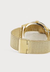 Bracelet de montre en maille dorée avec un fermoir lisse, présentant une finition métallique brillante. Des marquages visibles au dos de la montre incluent "Acier inoxydable."