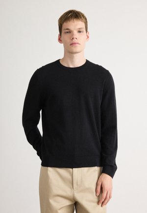 Pull en maille noir avec un col rond et des manches longues, présentant une texture lisse et un ourlet ajusté, associé à un pantalon beige.