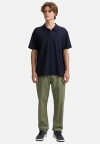 Navyblaues Polohemd mit Kragen und drei Knöpfen, kombiniert mit olivgrünen, lässig geschnittenen Hosen und grauen Sneakers. Einfaches Design, weicher Stoff.