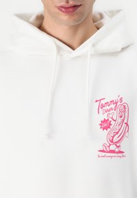 Witte hoodie met een roze grafiek van een cartoon-hotdog, inclusief de naam en slogan van het restaurant, met een trekkoordsluiting en een zachte textuur.