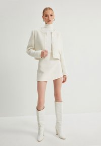 Hvid tweedjakke med rund hals og knaplukning, parret med en matchende kort nederdel. Hvide knæhøje støvler fuldender outfittet.