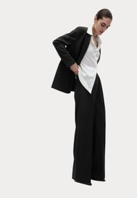 Schwarzer maßgeschneiderter Blazer über einem weißen Hemd, kombiniert mit weit geschnittenen schwarzen Hosen. Glattes Material, scharfer Kragen und elegante Linien sind hervorstechend.