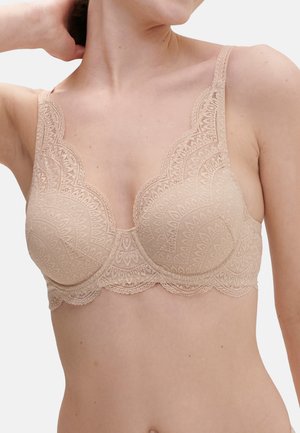 Kvinde iført en beige blonde-bh med bølget kant, der viser overkroppen og nakken, med den ene arm løftet bag hovedet.