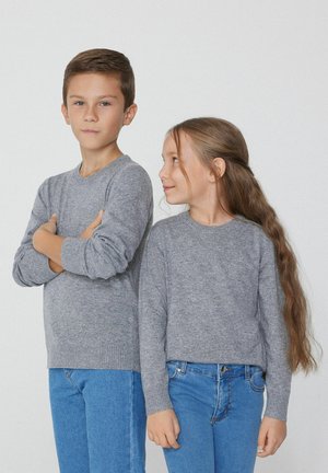 Graue Strickpullover mit gerippten Bündchen, kombiniert mit blauen Jeans. Der Junge steht mit verschränkten Armen, während das Mädchen zur Seite schaut.