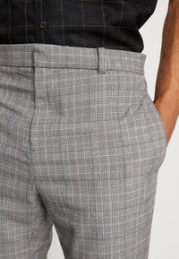 Pantalon à carreaux gris combinant plusieurs motifs de tartan, avec un tissu texturé et des poches latérales. La ceinture est plate avec des passants pour ceinture.