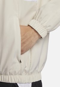 Giacca a vento beige chiaro con polsini e orlo elastici, dotata di tasca laterale e texture in tessuto leggera e liscia.