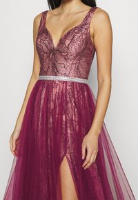 Burgundowa sukienka koronkowa z kwiatowym wzorem, głębokim dekoltem w kształcie V, przezroczystą narzutą oraz srebrnym paskiem z cekinami, z rozcięciem do uda i zwiewną sylwetką.