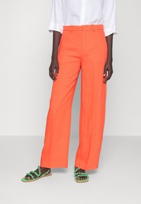 Pantaloni larghi in lino arancione con vita alta, abbinati a sandali neri con cinturini intrecciati verdi e suola piatta. Camicia bianca non dettagliata.