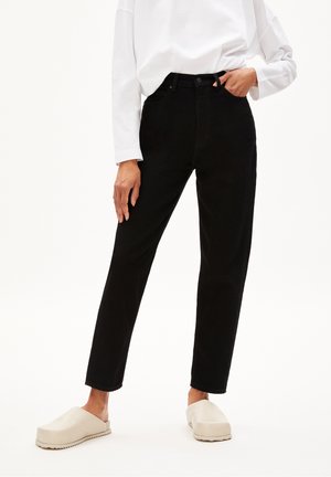 Frau trägt schwarze Jeans mit hohem Bund und geradem Bein, kombiniert mit weißem Langarmshirt und beigen Slippern, steht mit einer Hand in der Tasche.