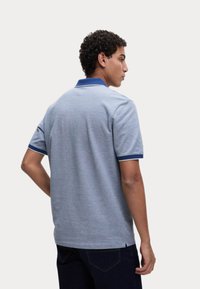 Polo shirt gris avec un col et des accents de manches bleus. Le tissu semble texturé, présentant une coupe classique et des fentes latérales pour le confort.