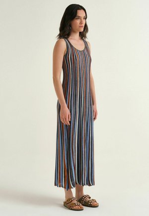 Donna in piedi che indossa un vestito senza maniche, lungo fino alla caviglia, a righe verticali blu, marroni, nere e bianche, abbinato a sandali decorati con borchie.