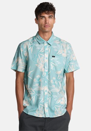 Chemise à manches courtes boutonnée en vert sarcelle clair avec un motif floral, présentant des feuilles blanches et beiges, et un petit logo brodé sur la poche poitrine.