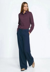 Bordowa koszula w grochy na guziki w połączeniu z granatowymi szerokimi spodniami. Beżowe buty z subtelnym wzorem, ręce w kieszeniach. Gładka tekstura tkaniny.