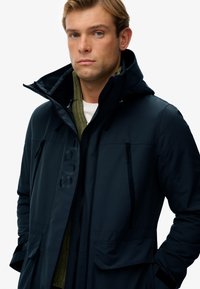 Marineblauwe hoodie-jack met ritszakken, textuurstof en "SUPER" merknaam op de voorkant. Gelaagd over een groene gebreide trui.