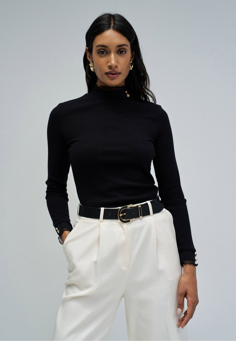 Woman wearing a black long-sleeve ribbed top with gold buttons, white high-waisted pants, black belt with gold buckle, and gold earrings.