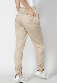 Pantalones jogger beige con banda elástica en la cintura, bolsillos laterales y traseros, tobillos ajustados y tejido suave. Combinados con zapatillas blancas y plateadas.