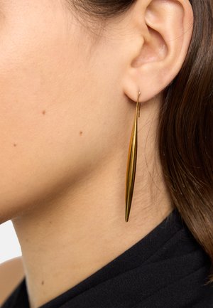 Zbliżenie na ucho kobiety noszącej długą, smukłą, złotą kolczyk wiszący na jasnej skórze i ciemnobrązowych włosach.