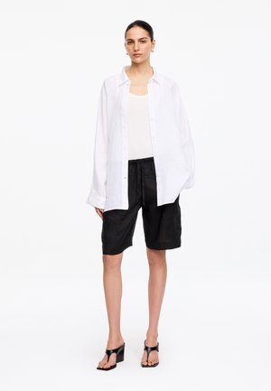 Kvinde iført en løs hvid skjorte med knapper over en hvid tanktop, sorte knælange shorts og sorte sandaler med hæl, stående på en hvid baggrund.