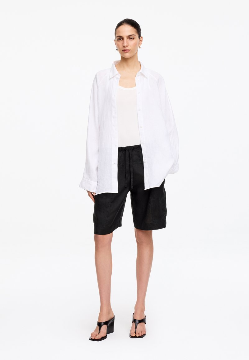 Femme portant une chemise blanche ample boutonnée sur un débardeur blanc, un short noir jusqu'aux genoux et des sandales noires à talons, debout sur un fond blanc.