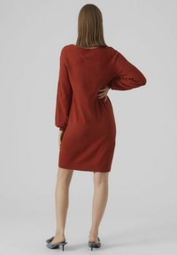 Robe-pull rouge à manches longues, avec un large décolleté et une texture douce. Elle arrive au-dessus du genou, associée à des mules à talons bleu clair.