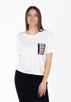 Uśmiechnięta kobieta w białej koszulce z czarną kieszonką ozdobioną powtarzającym się napisem „BLAH” i czerwonymi ustami, w połączeniu z czarną spódnicą.