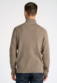 Maglione a coste marrone con collo alto, taglio aderente e maniche lunghe, abbinato a jeans blu scuro. Dettaglio del tessuto in rilievo visibile.